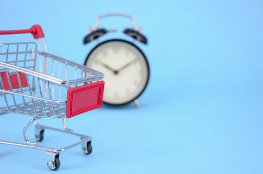 Shopping cart and classic alarm clock on blue background. Sale time market shop consumer concept. Selective focus.