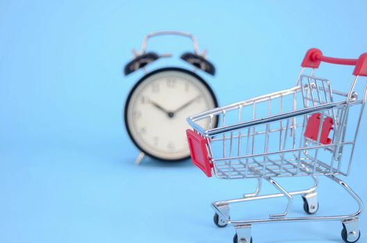 Shopping cart and classic alarm clock on blue background. Sale time market shop consumer concept. Selective focus.