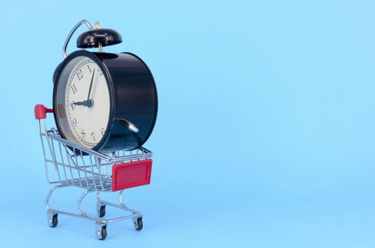 Shopping cart and classic alarm clock on blue background. Sale time market shop consumer concept. Selective focus.