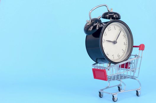 Shopping cart and classic alarm clock on blue background. Sale time market shop consumer concept. Selective focus.