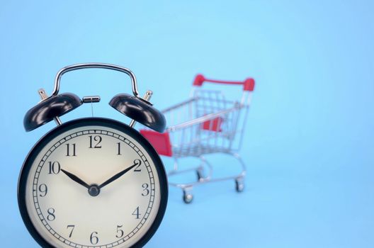 Shopping cart and classic alarm clock on blue background. Sale time market shop consumer concept. Selective focus.