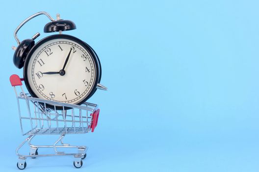 Shopping cart and classic alarm clock on blue background. Sale time market shop consumer concept. Selective focus.