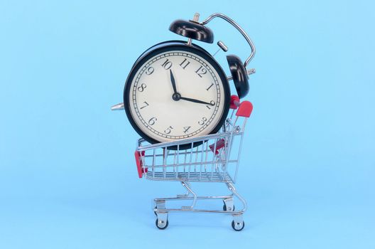 Shopping cart and classic alarm clock on blue background. Sale time market shop consumer concept. Selective focus.