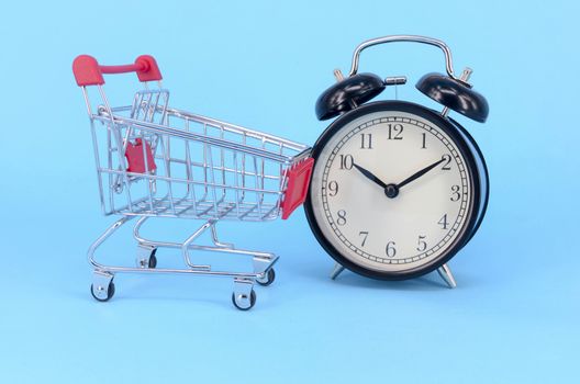 Shopping cart and classic alarm clock on blue background. Sale time market shop consumer concept. Selective focus.