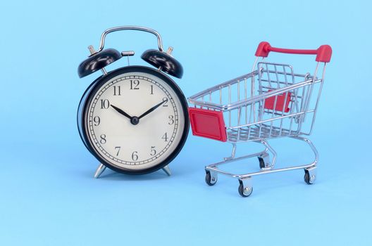 Shopping cart and classic alarm clock on blue background. Sale time market shop consumer concept. Selective focus.