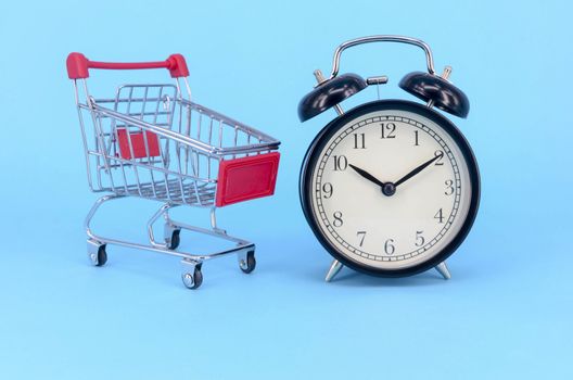 Shopping cart and classic alarm clock on blue background. Sale time market shop consumer concept. Selective focus.