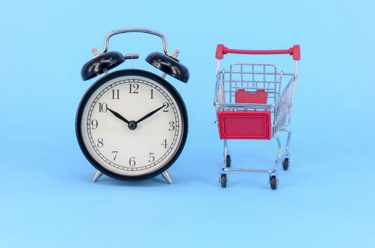 Shopping cart and classic alarm clock on blue background. Sale time market shop consumer concept. Selective focus.