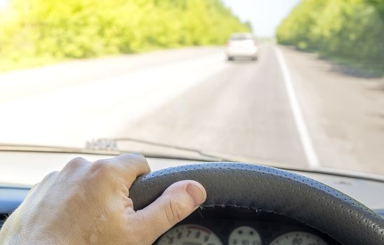 the driver hand on the steering wheel of a car that is passing on a country highway