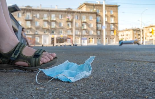 abandoned and used antibacterial medical mask lies on the asphalt at the feet of a man who is sitting on a bench in a city Park