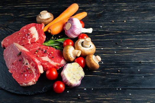 Three pieces of juicy raw beef with rosemary on a stone cutting board on a black wooden table background. Meat for barbecue or grill with vegetables pepper and salt seasoning