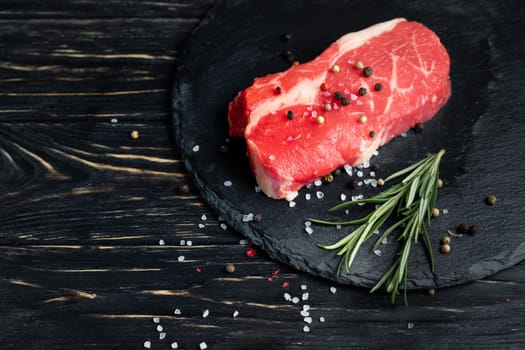 One pieces of juicy raw beef with rosemary on a stone cutting board on a black wooden table background. Meat for barbecue or grill sprinkled with pepper and salt seasoning