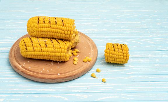 Grilled hot Corn cob lies on round cutting board plate blue wooden table background. Copy space for text.