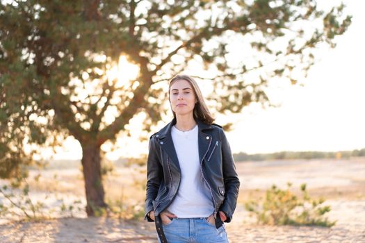 Young beautiful caucasian woman dressed leather jacket and jeans stands on the beach The rays of the sun break through the foliage of a tree background European girl in casual clothes walking outdoor