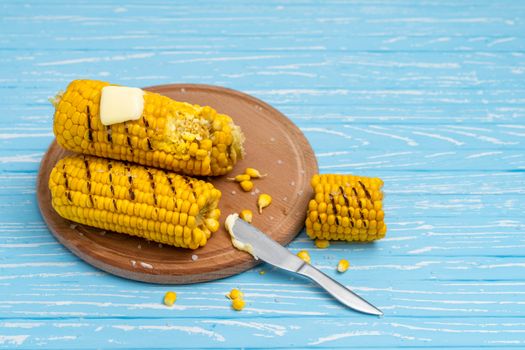 Grilled hot Corn cob lies on round cutting board plate blue wooden table background. Copy space for text.