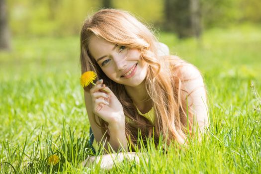 Happy smiling young woman laying on the green grass holding yellow dandelion flower
