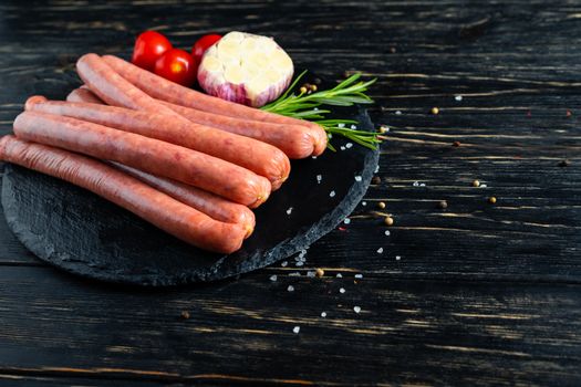 Juicy sausages for barbecue lie on a stone chopping board with rosemary garlic cherry tomatoes spices, pepper and coarse salt on the black wooden table background