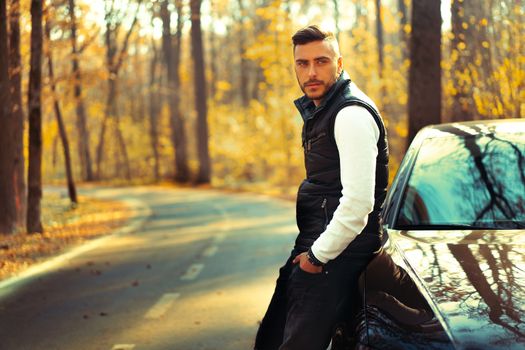 A handsome Caucasian man wearing style black vest standing near black car on nature. Young adult businessman travels by car on a beautiful sunny autumn day. Elite luxury driver.