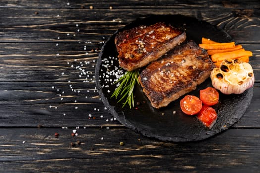 A juicy piece of fried meat with grilled cherry tomatoes garlic and carrot lies on a stone plate against a black wooden table. Degree of roasting well done. Food concept with copy space