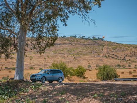 Gray SUV off-road vehicle parked under a Eucalyptus tree shade on a dirt road. The landscape of mountains and wheat fields with hay bales in the country area of Jordan Valley in northern Israel.