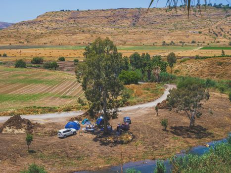 Landscape of country area of Jordan Valley in Israel with a group of people camping. SUV vehicles and tents under a tree near a river. Mountains and wheat fields with hay bales in the background.