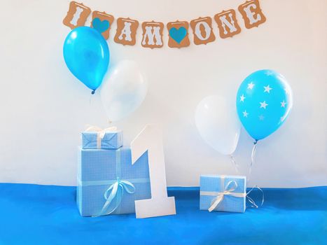 One year birthday decorations set. Decoration for a boy holiday party. Bunch of blue & white balloons, gift boxes, large white kappa board number one shape, and sign with the inscription "I am one".