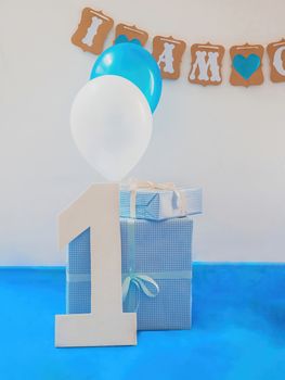 One year birthday decorations set. Decoration for a boy holiday party. Bunch of blue & white balloons, gift boxes, large white kappa board number one shape, and sign with the inscription "I am one".