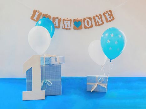 One year birthday decorations set. Decoration for a boy holiday party. Bunch of blue & white balloons, gift boxes, large white kappa board number one shape, and sign with the inscription "I am one".