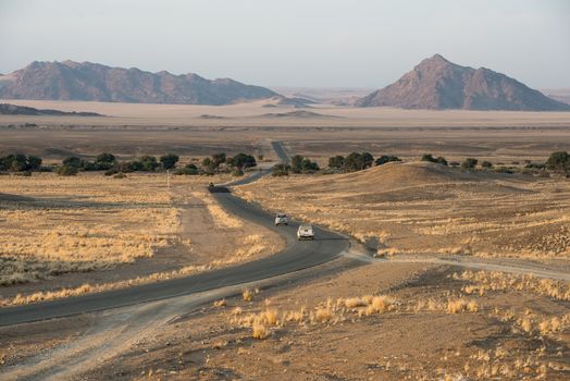 A modern asphalt road with tourist cars is bending across Namibian endless fields