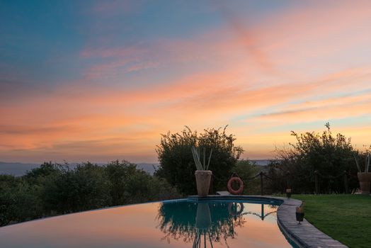 Morning colorful clouds and green Namibian trees are reflected in a mirror of a hotel swimming pool