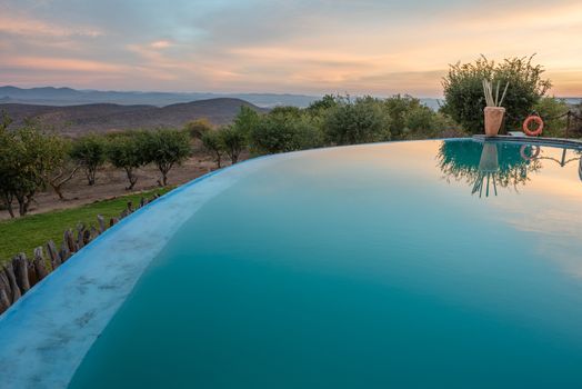 Morning colorful clouds and green Namibian trees are reflected in a mirror of a hotel swimming pool