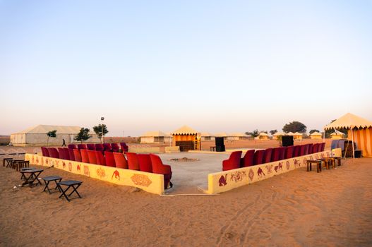 Wide angle shot of seating and entertainment area of a desert camp with sand around and tents in the distance. Shows the entertainment area in these adventure camps made for tourists and travellers