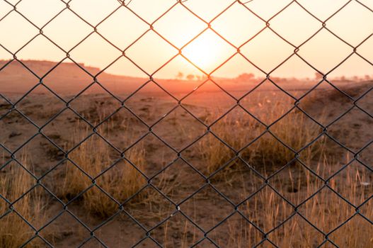 Chain link fence blocking the passage to beautiful sunset with barren desert with bushes and mountains in the distance. Shows the thar desert in India near jaisalmer sum with the aravalli range in teh distance