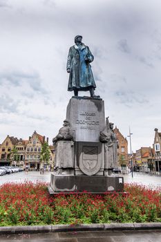 Diksmuide, Flanders, Belgium -  June 19, 2019: Grote Markt. Bronze statue of General Jacques de Dixmude on gray stone pedestal in red flowers against silver sky. Some Yellowish brown house facades.