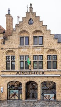 Diksmuide, Flanders, Belgium -  June 19, 2019: Grote Markt. Yellow brick facade with step gable of Pharmacy delplancke shows three bowed window with drug posters. Gray rainy sky.