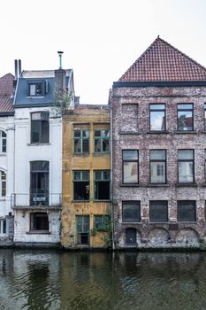 Gent, Flanders, Belgium -  June 21, 2019: Back facades of three houses built along and descending into the Leie River. Black greenish water, silver sky, white, yellow and pinkish stones with windows.