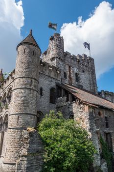 Gent, Flanders, Belgium -  June 21, 2019: Gray stone tower and other structures of Gravensteen, historic medieval castle of city against blue sky with white clouds. Flags on top, green foliage.