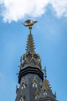 Gent, Flanders, Belgium -  June 21, 2019: Closeup of gulden draak, dragon statue, on top of dark stone Belfry clock tower against blue sky with white patches.