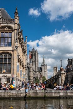 Gent, Flanders, Belgium -  June 21, 2019: School children and students enjoying sun sitting on quay of Leie River and Graslei. Three towers of city in back.. Blue sky and clouds.