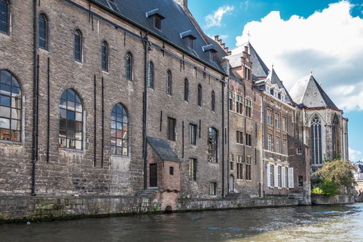 Gent, Flanders, Belgium -  June 21, 2019: Brown stone University building Het Pand bordering Leie River under blue-white cloudscape. Greenish water, Green foliage.