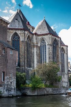Gent, Flanders, Belgium -  June 21, 2019: Brown gray stone building with gothic windows of Saint Michaels, Sint Michiels, church along Leie River under cloudscape. Green foliage.