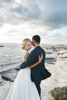 Happy couple hugging together on the beautiful landscape of the seacoast of Cyprus. Young woman in a wedding dress and groom in a suit posing on the cliff near the ocean. The concept of romance, travel, adventure.