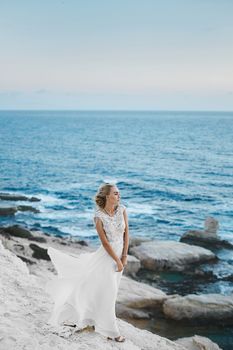 Young model woman with perfect body in stylish long white dress posing on the rocks on the seacoast at Cyprus.