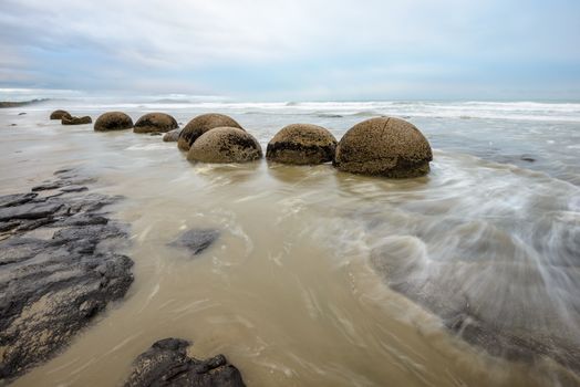 Impressive Moeraki boulders, cloudy morning sky and blurred Pacific Ocean waves at Moeraki Boulders Beach, New Zealand