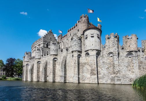 Gent, Flanders, Belgium -  June 21, 2019: Gray stone castle and ramparts of Gravensteen, historic medieval castle of city, behind its moad against blue sky with white clouds. Flags on top.