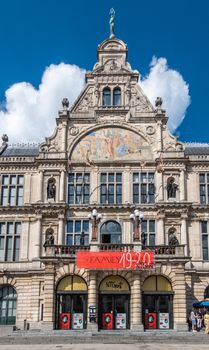 Gent, Flanders, Belgium -  June 21, 2019: Front facade of NTG Theater building on Sint-Baafsplein against blue sky with white clouds, Red and yellow advertisement on beige stone. Bronze statues.