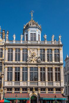 Brussels, Belgium - June 22, 2019: Beige stone facade and gable with statues on top of building named, Le Roy D’Espagne, with bar restaurant at ground, against blue sky.