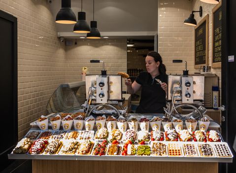 Brussels, Belgium - June 22, 2019: Open window with display of variety of Belgian waffles of small store has servant baking them, silver machines and white tiled background.