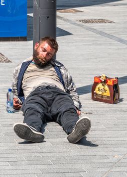 Brussels, Belgium - June 22, 2019: Filthy drunken lad sleeps in sun on gray stone Boulevard Anspach with six-pack of Leffe Abbey ale beers besides him.