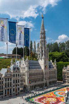 Brussels, Belgium - June 22, 2019: Mini-Europe exhibition park. Grand Place with flower carpet and City hall building with spire in miniature version against blue sky with white cloudscape. Green foliage.