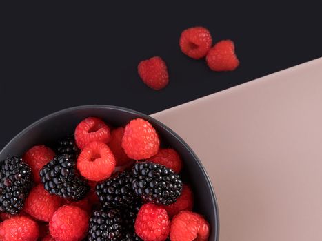 Blackberries and raspberries in a black bowl. Black and light pink background. Top view.